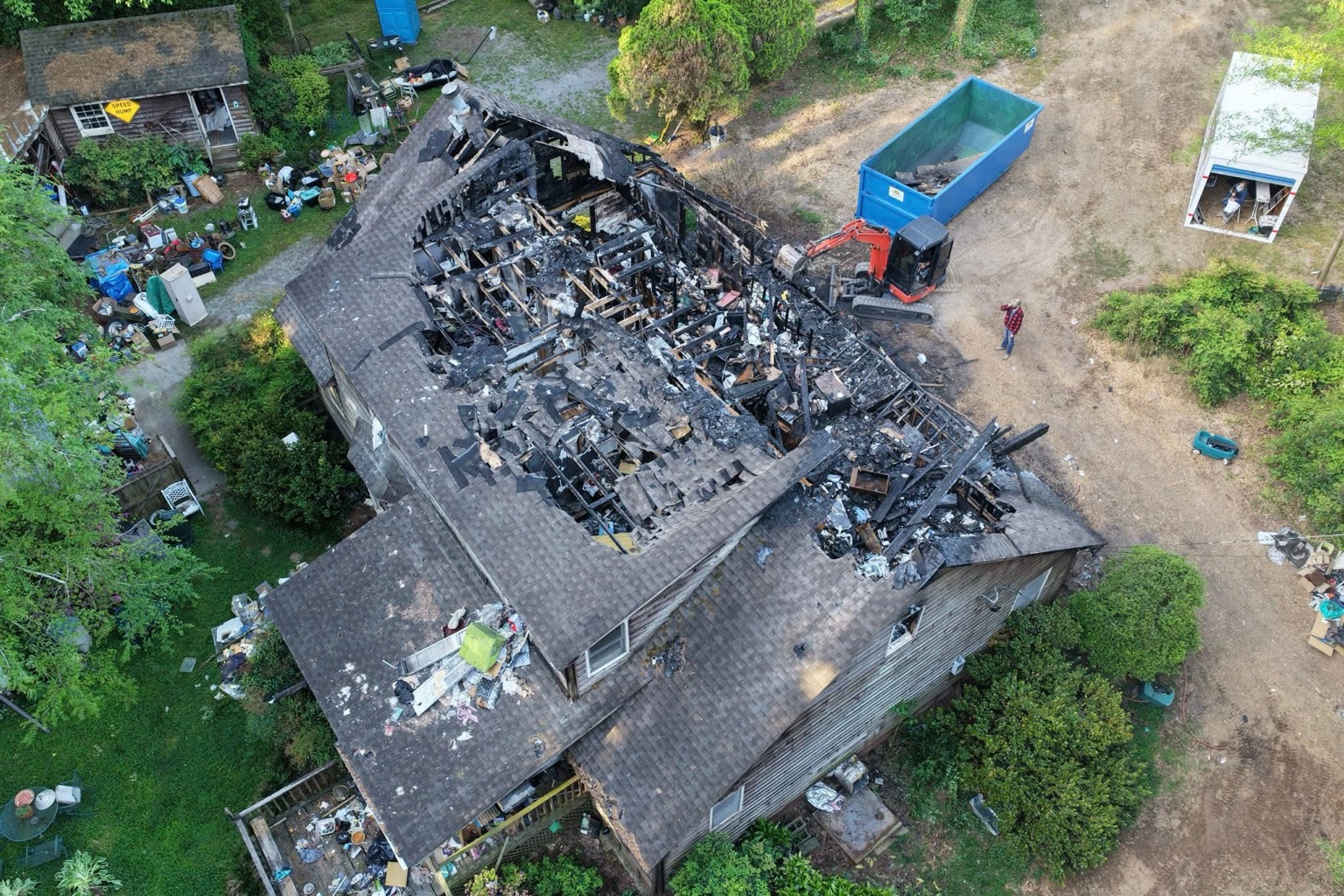 Aerial view of a fire-damaged house being demolished, with a blue dumpster and excavator on site